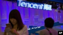 FILE - A woman uses her smartphone near a booth for the Chinese Internet company Tencent at the Global Mobile Internet Conference in Beijing, China, April 29, 2015.