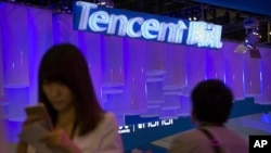 FILE - A woman uses her smartphone near a booth for the Chinese Internet company Tencent at the Global Mobile Internet Conference in Beijing, China, April 29, 2015.