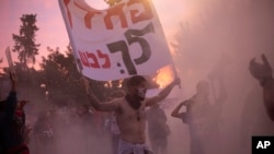 Pengunjuk rasa anti-pemerintah Israel memegang spanduk saat dia diselimuti asap dari api unggun di luar kediaman resmi PM Benjamin Netanyahu pada hari persidangan korupsinya semula dijadwalkan sebelum ditunda, di Yerusalem, 13 Januari 2021.(AP Photo/Maya Alleruzzo)