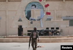 FILE - A Kurdish fighter from the People's Protection Units (YPG) carries his weapon as he walks at the faculty of economics where a defaced picture of Syrian President Bashar al-Assad is seen in the background, in the Ghwairan neighborhood of Hasaka, Syria, Aug. 22, 2016.