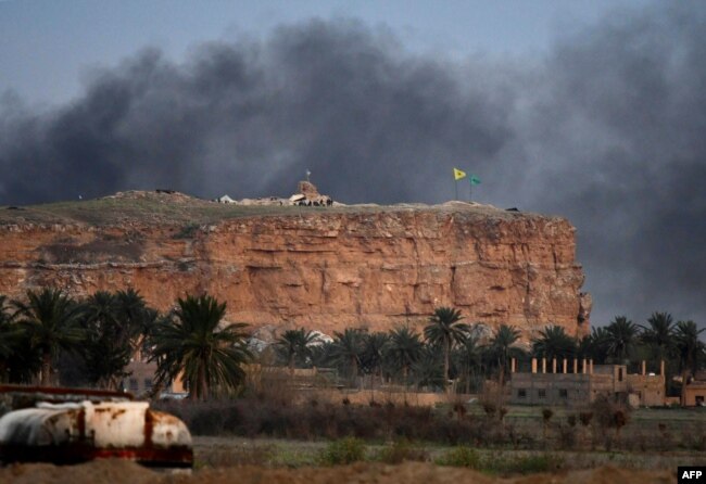 This picture taken on March 22, 2019 shows smoke rising over the village of Baghuz in the eastern Syrian province of Deir Ezzor, with the flags of the Kurdish People's Protection Units (Yellow) and Women's Protection Units (Green) seen flying.