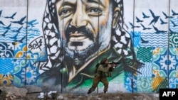 FILE - A member of the Israeli border guards looks through the scope of an assault rifle as he stands by a mural showing a graffiti image of late Palestinian leader Yasser Arafat, at the Qalandiya checkpoint near the West Bank city of Ramallah.
