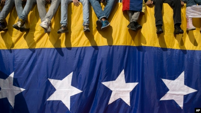 En esta foto la gente se sienta en una pared cubierta por una bandera venezolana durante una protesta contra el presidente Nicolás Maduro en Caracas, Venezuela.