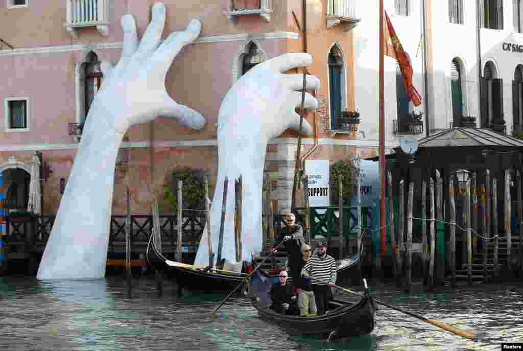 Para turis menaiki gondola di dekat karya seni karya seniman Italia Lorenzo Quinn di istana Ca&#39; Sagredo, Venesia, Italia.