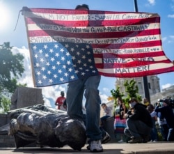 Gabriel Black Elk, who is Lakota, kneels on the neck of a fallen statue of Christopher Columbus and holds an American flag with the names of Native Americans killed by police, at the Minnesota state Capitol in St. Paul, Minn., June 10, 2020. (Evan Frost)