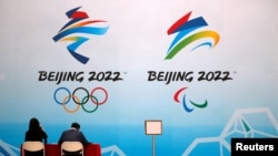 FILE - Staff members sit near a board with signs of the 2022 Olympic Winter Games, at the National Aquatics Center in Beijing, China, April 1, 2021.