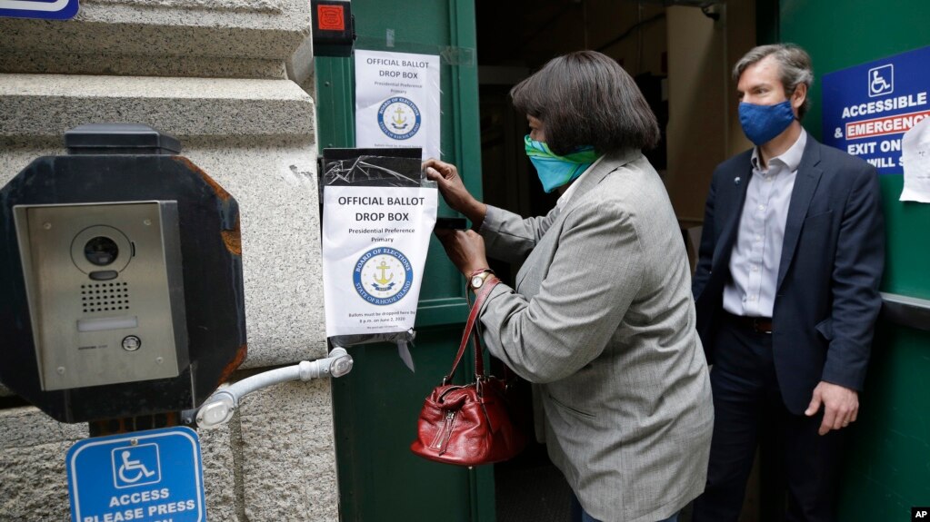 A voter, left, places a ballot in a secure box as Providence City Clerk Shawn Selleck, right, looks on, Tuesday, June 2, 2020, in Providence, R.I. Because so many voters asked for mail ballots, state electoral officials say mail ballots can be dropped off