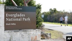 Visitors walk past a sign for Everglades National Park as they enter from overflow parking, Jan. 2, 2019, in Everglades National Park, Fla. Human feces, overflowing garbage, illegal off-roading and other damaging behavior were beginning to to overwhelm some of the national parks, as a partial government shutdown drags on.