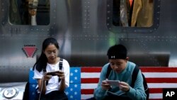 FILE - Shoppers sit on a bench with a decorated U.S. flag browsing their smartphones outside a fashion boutique selling U.S. brand clothing at the capital city's popular shopping mall in Beijing, Sept. 24, 2018.