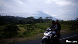 People ride a motorcycle past Mount Agung in the background, a volcano on the highest alert level, in Karangasem Regency, on the resort island of Bali, Indonesia, Sept. 24, 2017. 