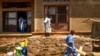 کارمند سردخانه ای در شهر بنی کنگو در حال ضدعفونی کردن ورودی سردخانه، و مرد دیگری که صلیب چوبی برای قبر یکی از کشته شدگان در دست حمل می کند.