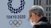 2020年3月24日一名戴着口罩的男子走过东京政府大楼上展示的2020年东京奥运会的徽标。