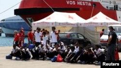 FILE - Migrants sit on the ground after disembarking from Vos Hestia ship of NGO "Save the Children" in the Sicilian harbor of Augusta, Italy, Aug. 4, 2017.