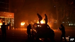 Demonstran berdiri di atas kendaraan sementara yang lainnya membakar kendaraan lain dalam sebuah kerusuhan yang meletus di Tripoli, Minggu (2/3).