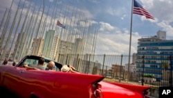 FILE - Tourists ride a vintage American convertible as they drive next to the American flag and a Cuban flag at the United States Embassy in Havana, Cuba, March 18, 2016. AT&T has reached a deal for enhanced roaming and other cellphone services for wireless customers visiting Cuba.