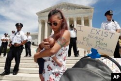 As security guards the steps of the Supreme Court, Kristin Mink of Silver Spring, Md., holds her three-week-old daughter by a sign she brought that says, "I exist because my Mom had an abortion," as Mink joined a protest against abortion bans, May 21, 201
