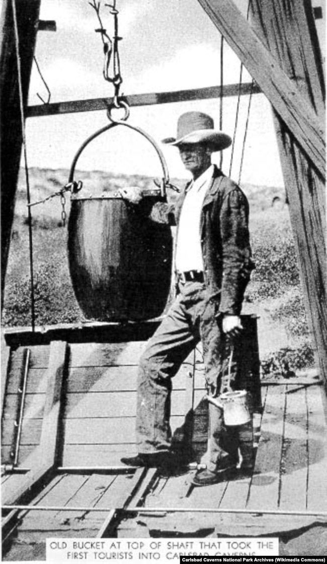 James Larkin "Jim" White standing next to a guano bucket atop the guano shaft at Carlsbad Caverns. The bucket was used to carry the first tourists into the caverns.