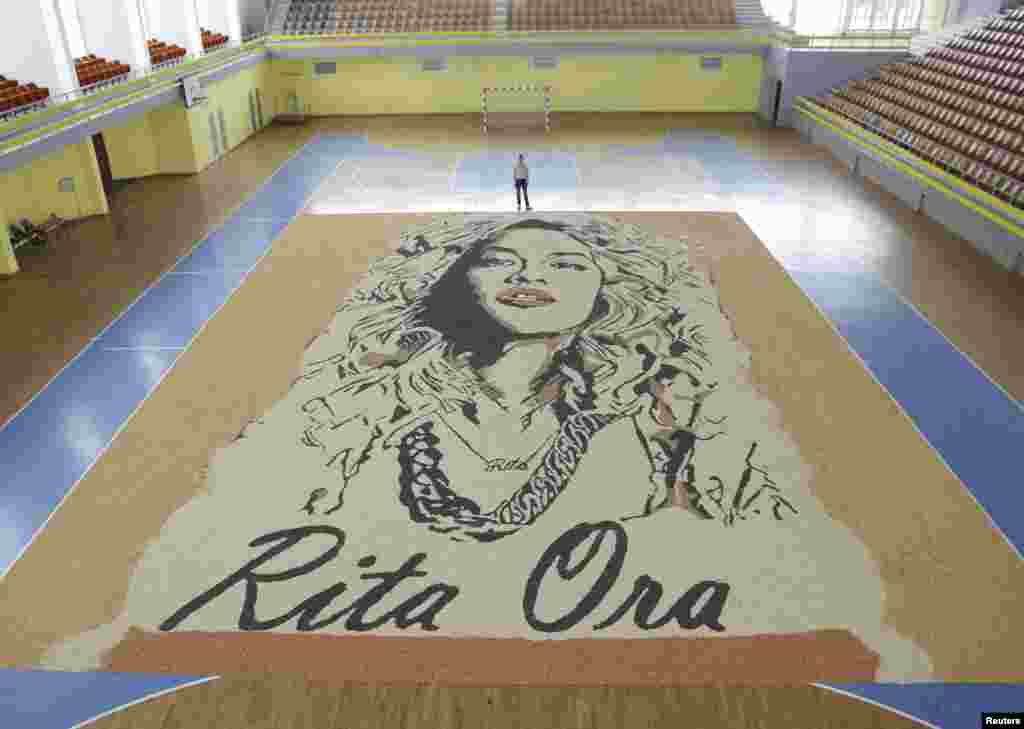 សិល្បករ​ជនជាតិ​កូសូវ៉ូ​ឈរ​ថតរូប​នៅ​ក្បែរផ្ទាំងគំនូរ​តារាចម្រៀង​ស្រី​ជនជាតិ​អាល់បានី​ អ្នកនាង Rita Ora, ដែល​លោក​បាន​ធ្វើ​ឡើង​ពី​គ្រាប់​រុក្ខជាតិ​ក្នុង​ទីក្រុង​ Gjakova។