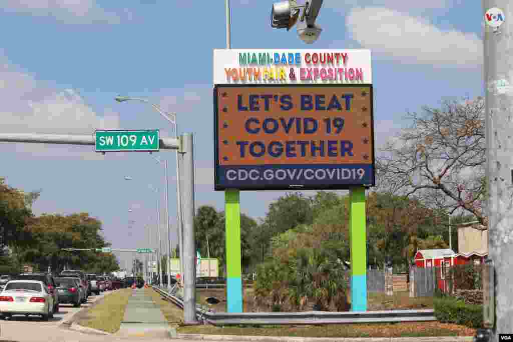 Un cartel anima: &quot;Vamos a vencer juntos al (nuevo coronavirus) COVID-19&quot;. Una larga fila de autos bloquea una entrada de la feria de la Juventud en Miami-Dade, donde el viernes se distribuyeron alimentos gratis a personas necesitadas. [Foto: Luis F. Rojas/Voa Noticias].