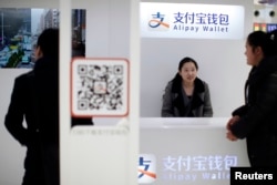 Nhân viên hỗ trợ bán hàng đứng bên dưới logo của Alipay tại một nhà ga ở Thượng Hải ngày 9/2/2015.