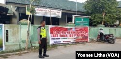 Seorang petugas Polisi Lalu Lintas Polres Palu berdiri di depan sebuah baliho yang dipasang di pagar Kantor Camat Tatanga yang bertuliskan ajakan mewujudkan Tatanga bersinar, bersih dari narkoba, Jumat, 26 Juli 2019. (Foto: Yoanes Litha/VOA)