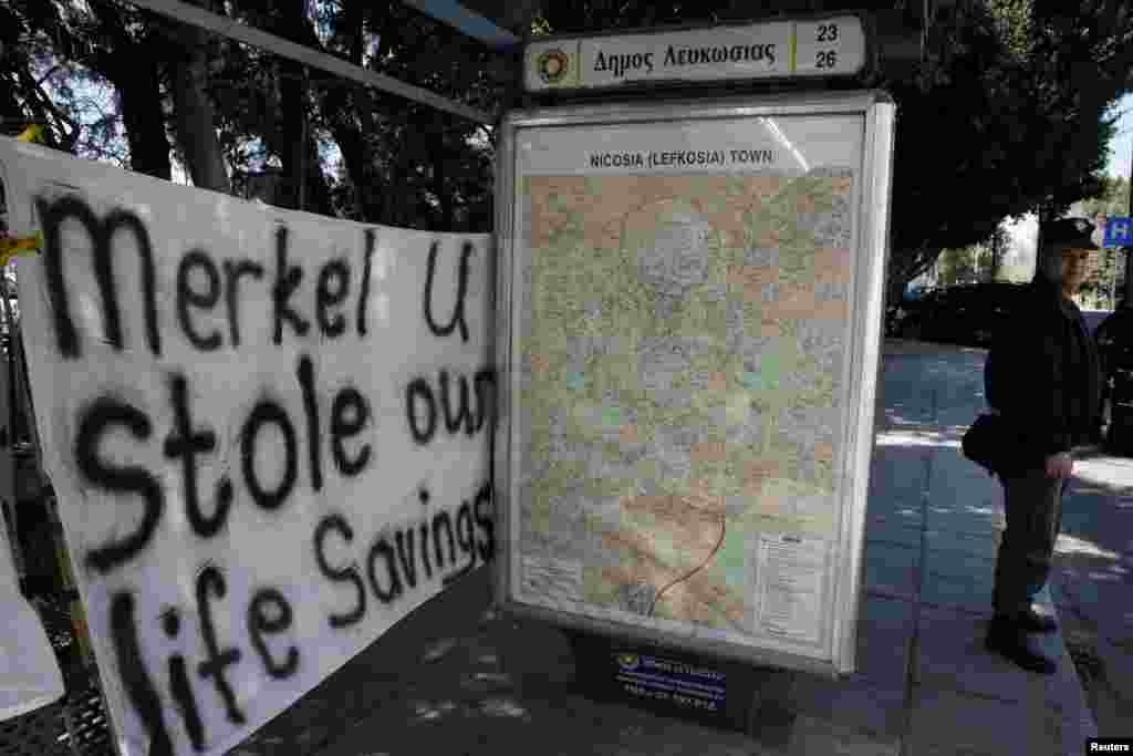 一名警察2013年3月18日站在塞浦路斯尼科西亚一回附近的一个公共汽车站前面。而一个反对获得国际紧急援助资金的标语牌就摆放在议会大楼的外面。