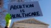 Žena u liječničkoj uniformi drži poster na kojem piše "Abortus je zdravstvena zaštita" dok zagovornici prava na abortus i demonstranti protiv abortusa demonstriraju ispred Vrhovnog suda SAD-a, srijeda, 1. decembra 2021., u Washingtonu. (AP Photo/Andrew Harnik)
