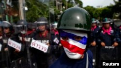 FILE - An anti-government protester wearing a mask painted in the colors of the Thai national flag looks on as riot police officers stand guard outside the parliament in Bangkok, Aug. 7, 2013. 