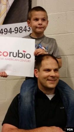 Mark Jones of Summerville, whose son Rig is perched on his shoulders, holds a Marco Rubio sign at a South Carolina rally Feb. 19, 2016. Even after the rally, Jones says he's still undecided, though "90 percent" for Rubio. (C. Guensburg/VOA)