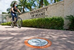 A cyclist, wearing a mask to prevent the spread of the new coronavirus, rides by an entrance to the University of Miami, Tuesday, Aug. 25, 2020, in Coral Gables, Fla. (AP Photo/Wilfredo Lee)