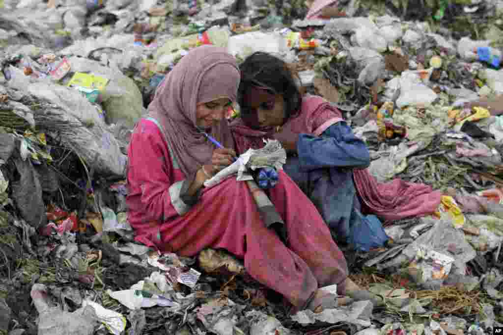 Seorang anak perempuan Pakistan yang menjadi pemulung menulis di buku yang ia ambil dari tumpukan sampah, sementara seorang anak perempuan lainnya duduk di sebelahnya di Lahore, Pakistan.