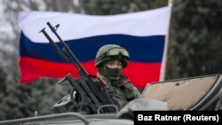 Seorang pria pro-Rusia (tidak terlihat) memegang bendera Rusia di belakang seorang prajurit bersenjata di atas kendaraan tentara Rusia di luar pos penjaga perbatasan Ukraina di kota Balaclava, Krimea, 1 Maret 2014. (Foto: REUTERS/Baz Ratner)