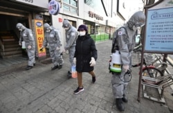 A woman wearing a face mask walks past South Korean soldiers wearing protective gear as they spray disinfectant on the street to help prevent the spread of the COVID-19 coronavirus, in Seoul on March 6, 2020.