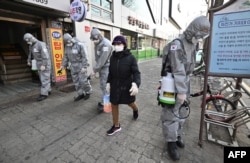 A woman wearing a face mask walks past South Korean soldiers wearing protective gear as they spray disinfectant on the street to help prevent the spread of the COVID-19 coronavirus, in Seoul on March 6, 2020.