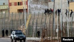 FILE - African migrants are seen sitting atop a border fence as Spanish Civil Guard officers stand below, on the border between Morocco and Spain's north African enclave of Melilla February 10, 2015.