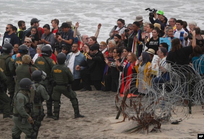 U.S. police and Border Patrol hold a line as members of an inter-faith group, showing support for Central American asylum-seekers who arrived in recent caravans and calling for an end to detaining and deporting immigrants, pray during a protest in San Diego, as seen from across the border wall in Tijuana, Mexico, Monday, Dec. 10, 2018.