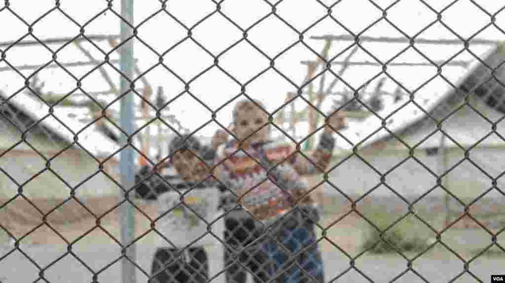 Anak-anak mengintip dari pagar kawat kamp Schisto. Para pengungsi bebas keluar masuk kamp, tapi banyak yang merasa terperangkap akibat proses lambat dan tidak pasti dari relokasi mereka ke tempat lain di Eropa, atau mencari suaka di Yunani. (VOA/J. Owens)