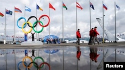 People walk past as the Olympic rings and the cauldron for the Olympic flame are reflected in a puddle of water on the Olympic Park as preparations continue for the Sochi 2014 Winter Olympics, Jan. 30, 2014.