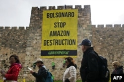 Para wisatawan berjalan melewati para aktivis Greenpeace yang sedang menggantungkan spanduk besar pada pembatas Kota Tua Yerusalem dengan pesan mengenai hutan Amazon untuk Presiden Brasil yang sedang berkunjung ke Yerusalem, 1 April 2019. (Foto: AFP)