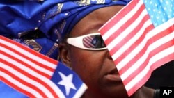 FILE - A market vendor stands among a group of women waving Liberian and American flags to show support for visiting U.S. Secretary of State Hillary Rodham Clinton, in Monrovia, Liberia. Obama recently lifted US sanctions on Liberia.