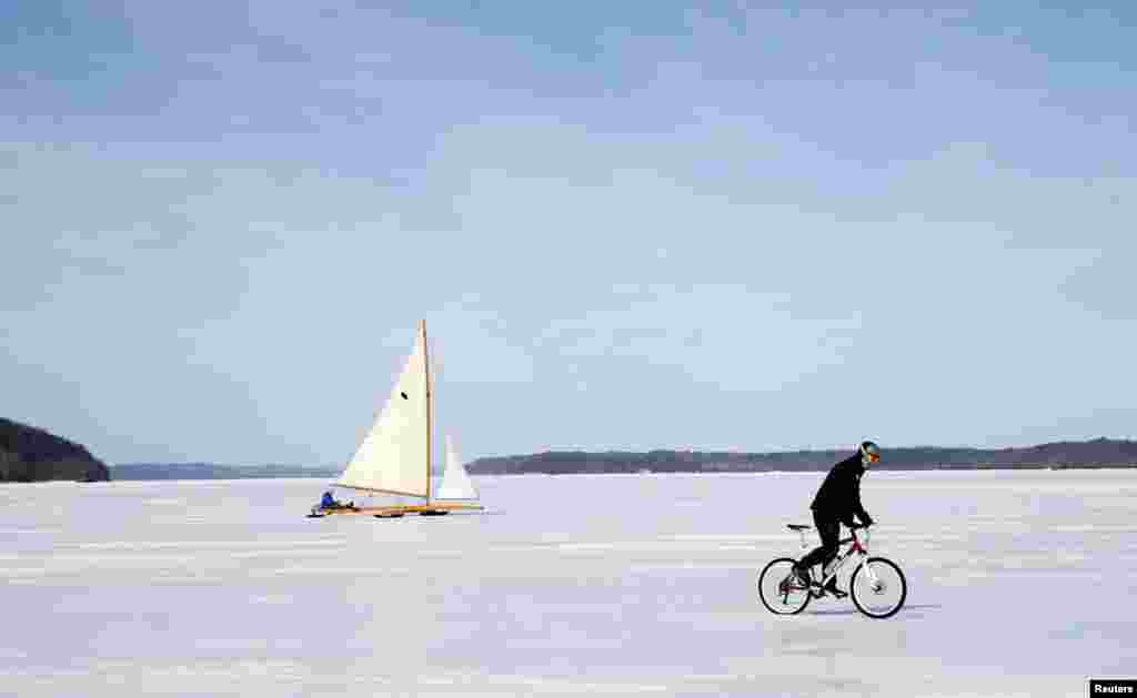 Một chiếc thuyền kiểu cổ lướt trên băng phía sau một người đi xe đạp vượt qua Sông Hudson đóng băng gần Astor Point tại Barrytown, New York, ngày 7 tháng 3, 2014. Những người đi thuyền lướt băng thuộc Câu lạc bộ thuyền buồm lướt băng trên Sông Hudson tận hưởng một trong những mùa lướt băng bằng thuyền tốt nhất và dài nhất trong những năm gần đây, khi nhiệt độ Bắc cực ảnh hưởng đến mùa đông tại vùng Đông Bắc nước Mỹ làm nước đóng băng dày cho phép họ lướt những con thuyền kiểu cổ bằng gỗ nặng nề.