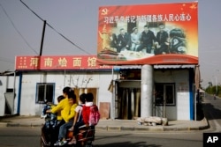 Seorang peremmpuan Uighur sedang mengendarai skuter bersama anak-anaknya melewati papan iklan bergambar Presiden China Xi Jinping bersama sejumlah tetua suku Uighur di Hotan, Provinsi Xinjiang, 23 Maret 2021. (Foto: Andy Wong/AP Photo)