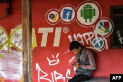 FILE - A man plays with his mobile phone in front of a cellular provider's advertisement with a Facebook logo in Jakarta, March 22, 2018.