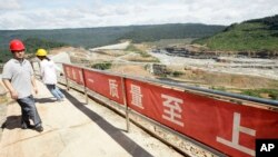 A Chinese engineer, left, walks by a fence with Chinese slogans reading: Safety first, highest quality and quantity near an entrance of a dam construction site by China National Heavy Machinery Corporation on the Tatay River in Koh Kong province, file photo. 