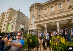 Fernando Rodriguez, left, uses his cellphone to take a selfie as workers from Eastern Shores Flagpoles raise a flagpole at the Cuban Interest Section in Washington in preparation for re-opening of embassies in Havana and Washington, June 10, 2015.