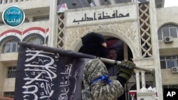 FILE - In this photo posted on the Twitter page of Syria's al-Qaida-linked Nusra Front, March 28, 2015, a fighter from Syria's al-Qaida-linked Nusra Front holds his group flag as he stands in front of the governor building in Idlib province, Syria. 