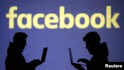 Silhouettes of laptop users are seen next to a screen projection of Facebook logo in this picture illustration taken March 28, 2018. 