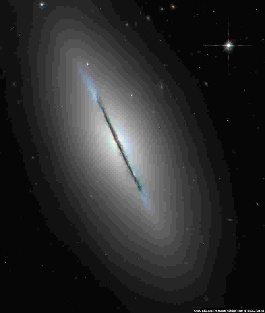 На этом снимке галактики NGC 5866 (также известной как &laquo;Веретено&raquo;) можно подробно рассмотреть галактику с необычного ракурса - с ребра. Вдоль плоскости галактики четко различимы темные области космической пыли. В центре, вокруг светящегося галактического ядра можно заметить едва различимое красное сияние - это балдж, скопление плотно расположенных старых звезд.