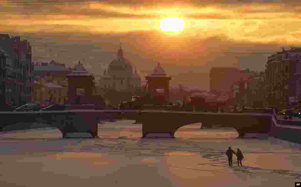 A couple walk on the ice on the frozen Fontanka River at sunset in St. Petersburg, Russia, with Troitsky (Trinity) Cathedral in the background.