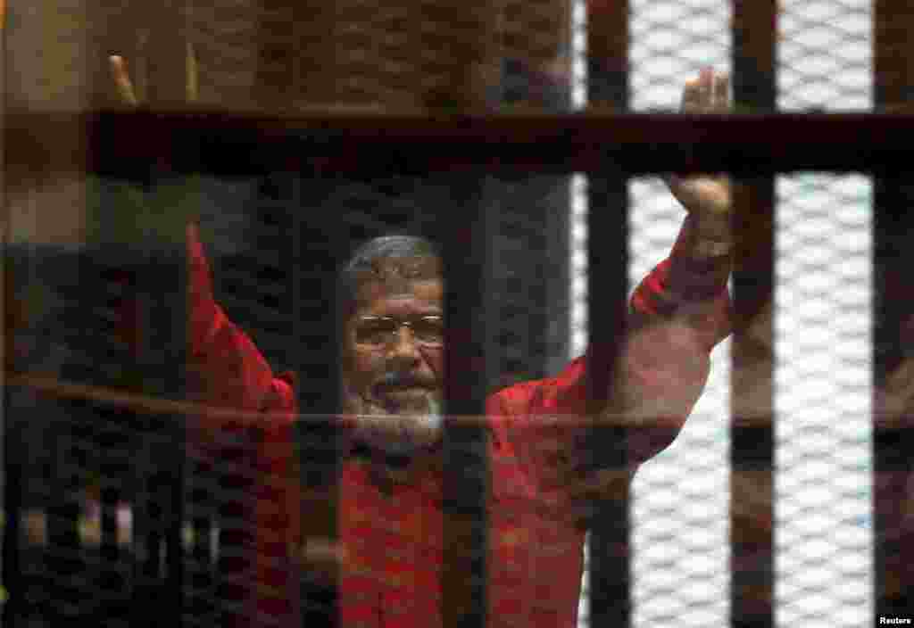 L'ancien président Mohamed Mursi salue ses avocats et des personnes derrière les barrières à la cour, portant un uniforme des prisonniers condamné à mort, lors de son procès le 21 juin 2015 au Caire en Egypte. Lire l'article ici.